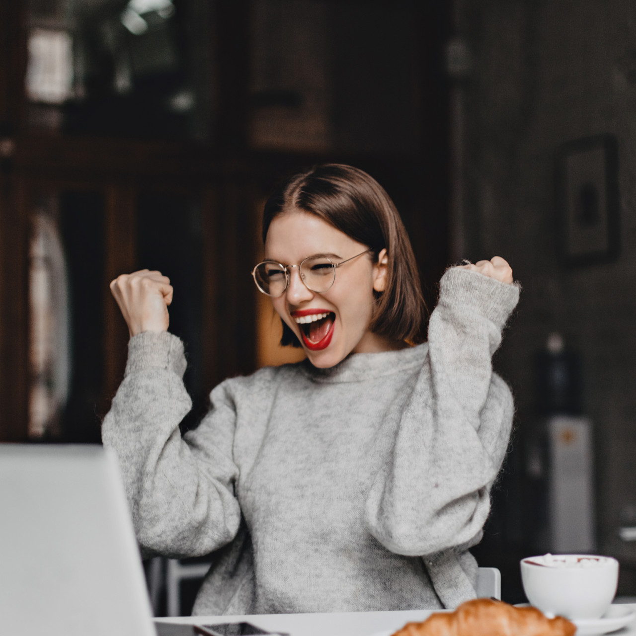 Photo of an excited young woman cheering in front of laptop at a coffee shop.