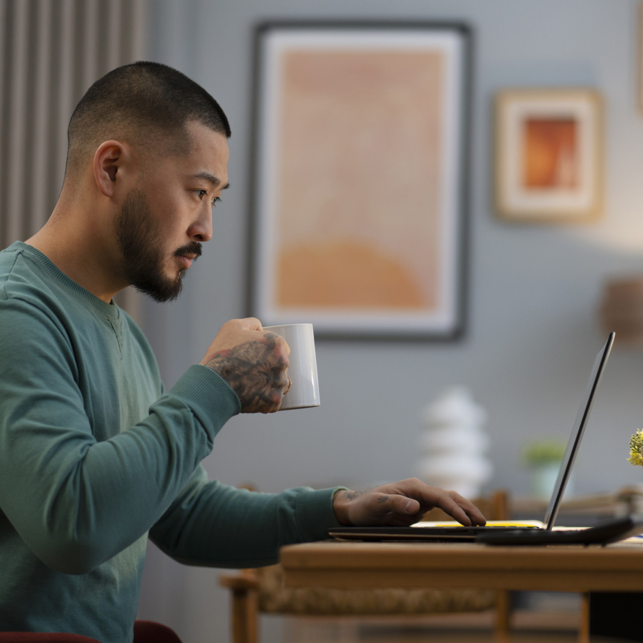 Photo of a man holding a coffee mug working on his laptop at home.