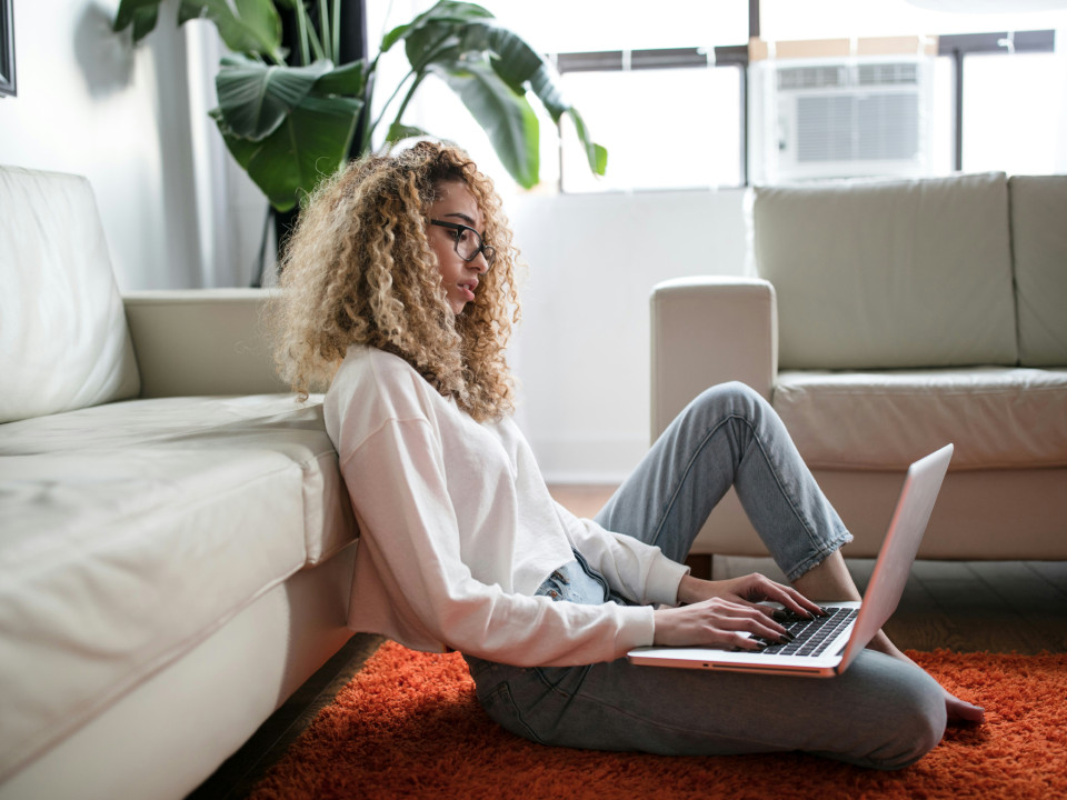 Photo of a young woman working on her laptop sitting on the floor in the living room.