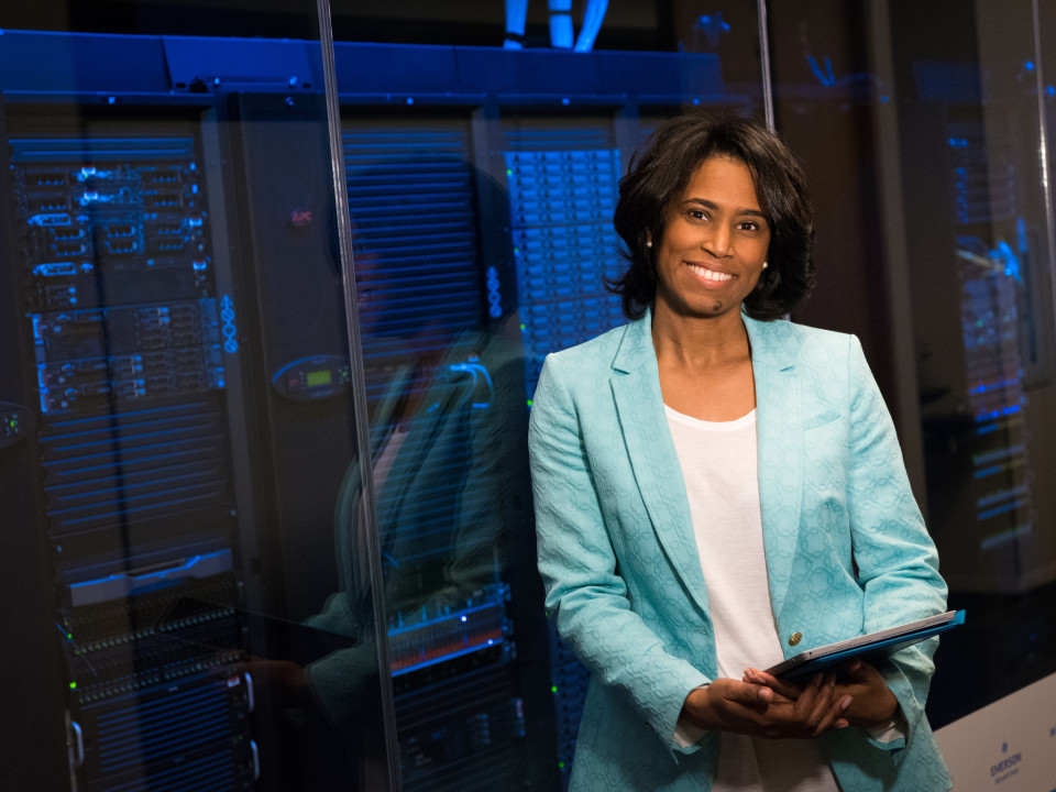 Photo of a woman holding a tablet standing beside server racks.